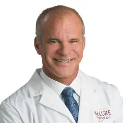 Man in a white medical coat with "Allure" logo, smiling, against a white background.