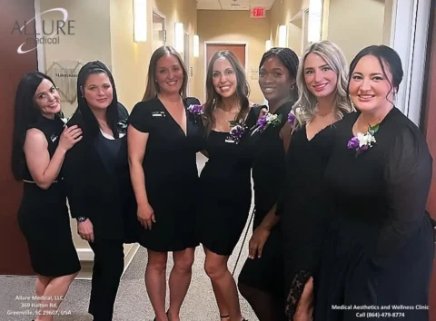 A group of seven women in black outfits stand in a hallway, smiling at the camera. The background features a wall with "Allure Medical" signage, and the women have name tags and purple flower accessories.