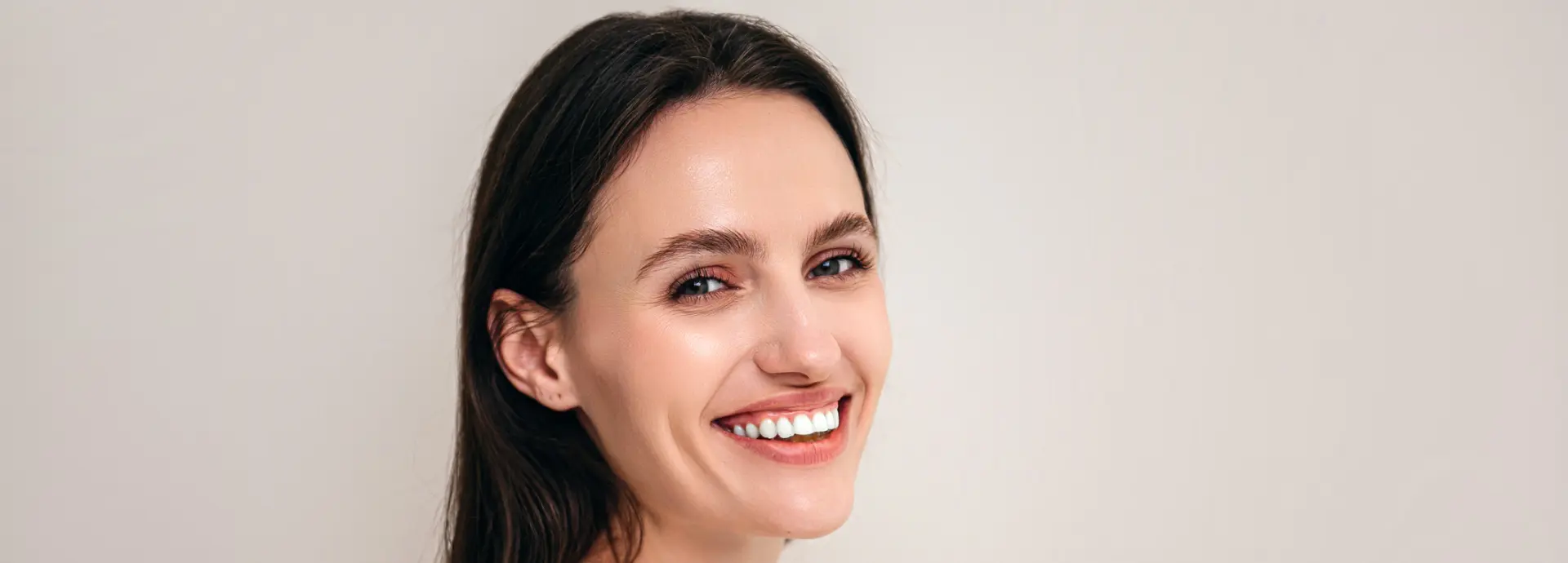 Close-up of a woman with long dark hair and light skin, smiling warmly at the camera against a plain background.