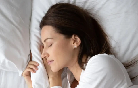 A woman with long dark hair sleeps peacefully on her side, resting her head on a white pillow and wearing a white top.